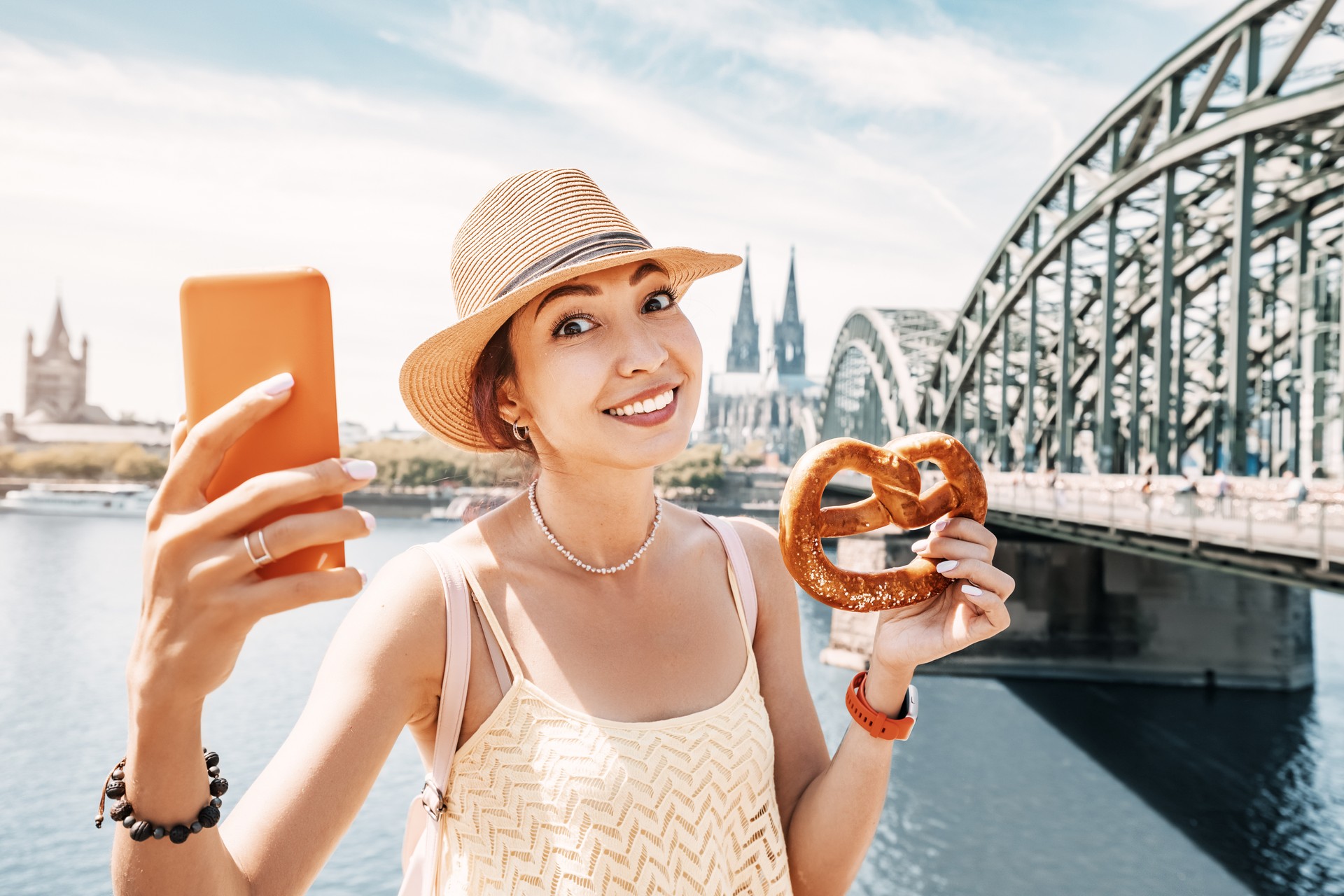 Asiatisches Touristenmädchen beim Selfie-Foto mit Brezel und Kölner Domgebäude und Rheinbrücke im Hintergrund. Reisen und Tourismus in Deutschland