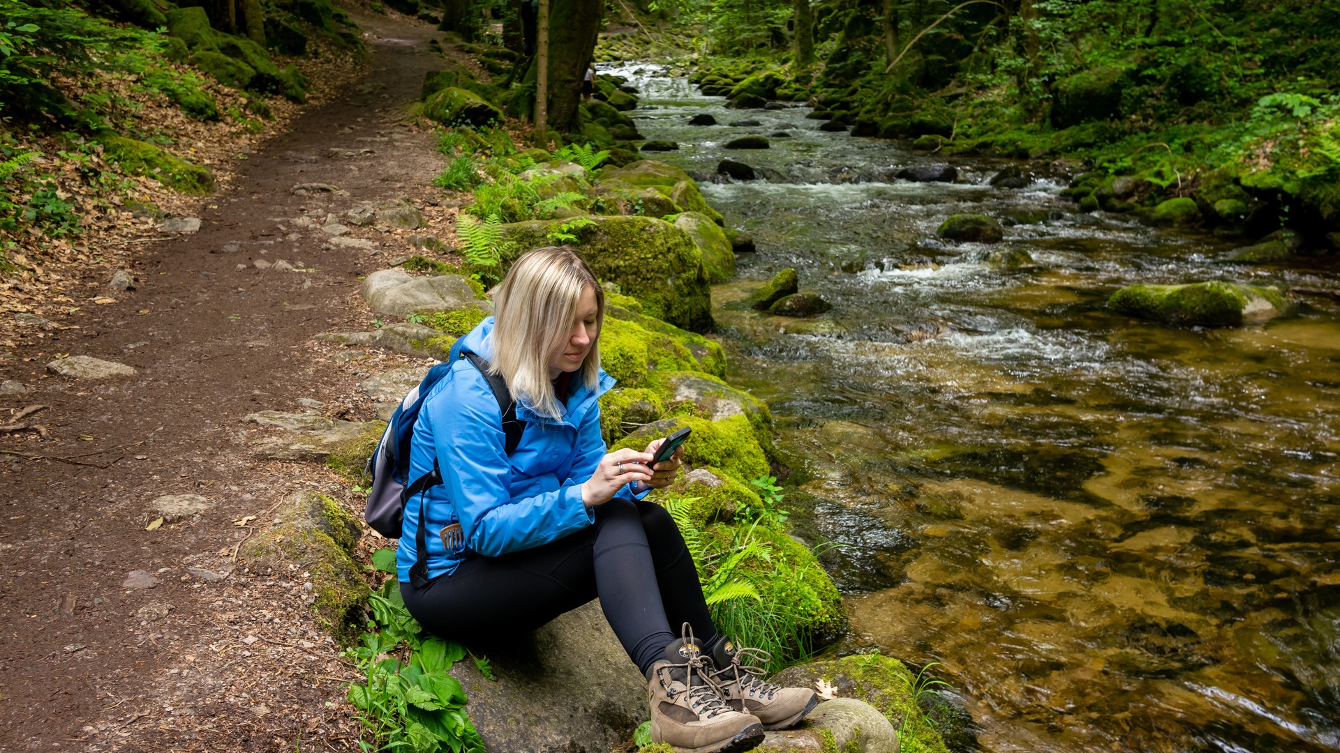 Junge blonde Frau in blauem Parka und Wanderschuhen mit Smartphone.  Wanderausflug.